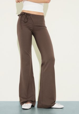 Nohy v hnedých zvonových nohaviciach s detailom na viazanie v páse a biele ploché topánky, stojace na svetlom pozadí.