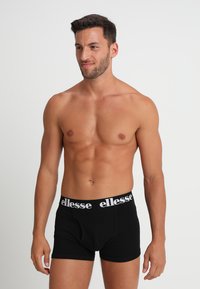 Svarta boxertrosor med en vit logoband, i bomullsmaterial, figursydd design och slät textur, bärs av en stående manlig modell.