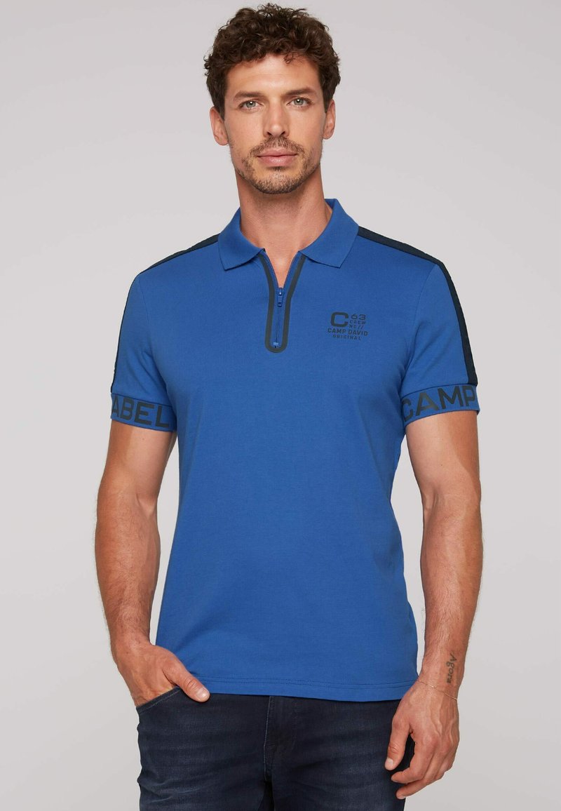 Camisa polo azul con cuello de cremallera, con detalles en negro en los hombros y los puños de las mangas. Incluye un logo impreso en el pecho y en la manga.