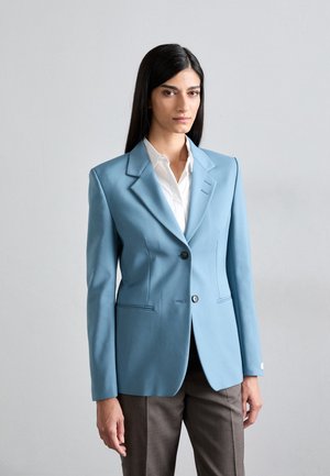 Light blue tailored blazer with structured shoulders, single button closure, and two front pockets. Paired with a white shirt and brown trousers.