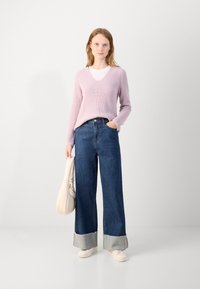 Pulover tricotat de culoare violet deschis cu decolteu în V, asociat cu blugi din denim albastri cu picioare largi și tiv gri. Modelul ține o geantă de culoare crem.