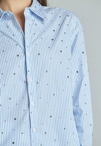 Camicia a righe blu chiaro con piccoli perni d'argento, chiusura a bottoni e design classico del colletto. Tessuto testurizzato con finitura liscia.