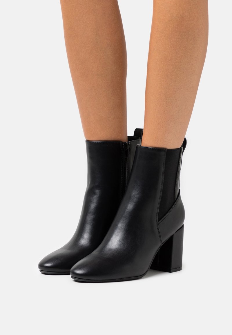 Jambes portant des bottines en cuir noir à talons blocs et fermetures éclair latérales, posées sur un fond clair uni.