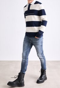 Sweter w paski w kolorze granatowym i kremowym z kołnierzem polo, z logo na piersi, w połączeniu z niebieskimi dżinsami i czarnymi, wiązanymi butami.