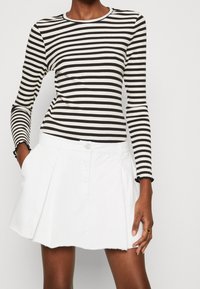 Czarna i biała bluzka w paski z długim rękawem o prążkowanej fakturze, zestawiona z białą spódnicą ozdobioną guzikiem i plisami. Detal z postrzępionym brzegiem.