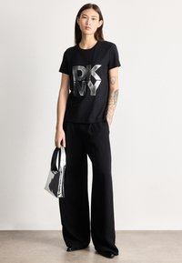 Jovem mulher com cabelo castanho escuro comprido, vestindo t-shirt preta DKNY, calças pretas de perna larga, segurando uma mala prateada e exibindo tatuagens no braço direito.