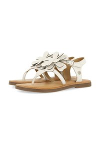 Sandalias blancas con un diseño floral hecho de pétalos superpuestos. Presentan una suela plana y una correa ajustable en el tobillo. Textura de cuero suave.