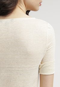 Jasno beżowa bluzka z krótkim rękawem, o fakturowanym, paskowanym wzorze oraz okrągłym dekoltem. Materiał wydaje się miękki i lekki.