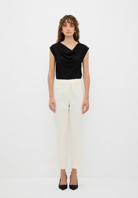Zwarte gedrapeerde top met korte mouwen, gecombineerd met crème op maat gemaakte broek en zwarte puntige hakken, tegen een effen witte achtergrond.