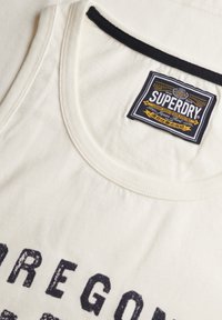Wit mouwloos shirt met zwarte halslijn en vetgedrukte tekst "OREGON" in donkere letters. Kenmerkt zich door een Superdry-label op de binnenkant van de hals.