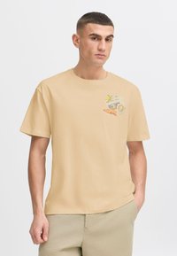 Short-sleeve, beige cotton t-shirt featuring a small graphic of fruits and a newspaper-style design printed on the left chest area.