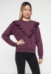 Sweatshirt violet avec un ourlet ondulé en forme de V sur le devant, à manches longues et avec un ourlet côtelé. Assorti à un pantalon noir.