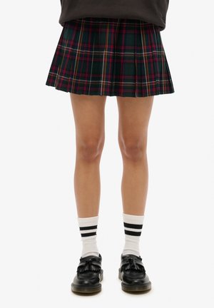 Jambes portant une jupe plissée à carreaux vert foncé et rouge, des chaussettes blanches avec deux bandes noires et des mocassins noirs à pompons.