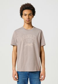 Jasnobrązowa bawełniana koszulka T-shirt z wyblakłym nadrukiem skaczącego pantery i szachownic. Krótkie rękawy i okrągły dekolt.