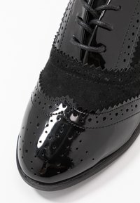 Chaussures noires avec un mélange de cuir verni et de daim, présentant des détails de brogue élaborés, un bout rond et des lacets noirs.