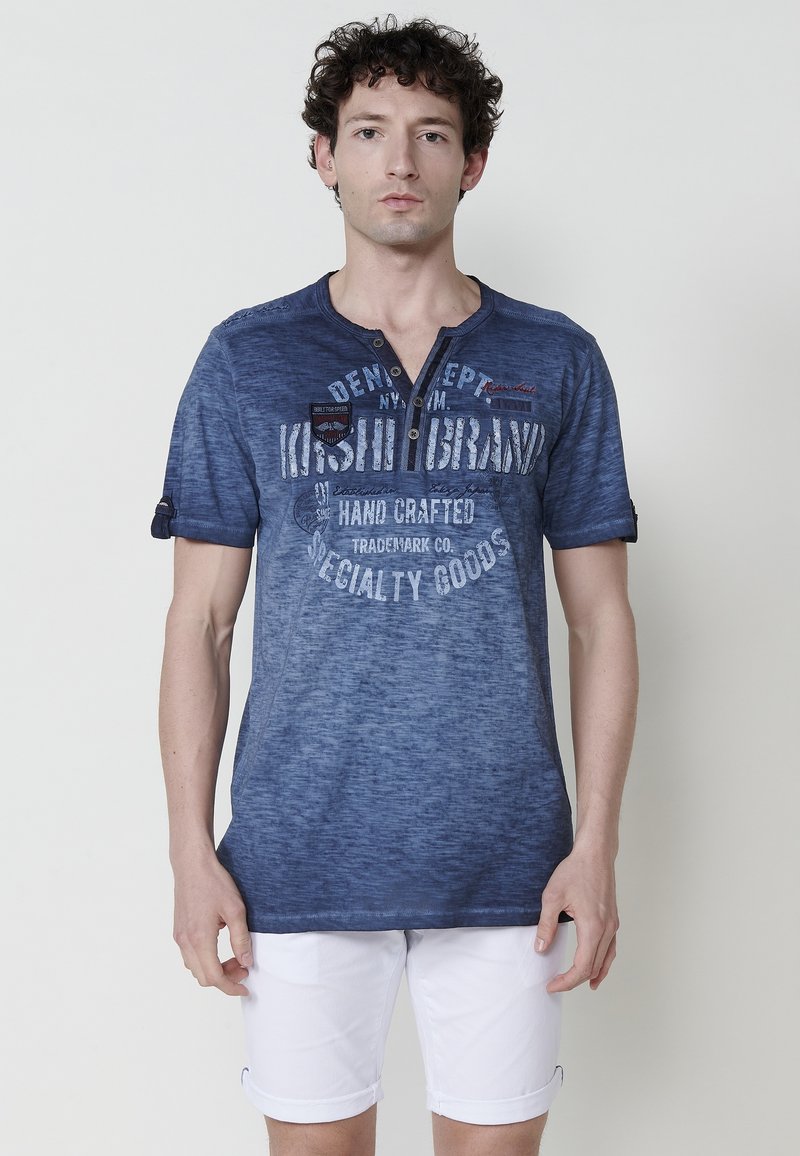 Laivansinisestä puuvillasta valmistettu t-paita, jossa on V-pääntie, lyhyet hihat ja koristeellisia tekstejä ja logoja. Ominaisuuksina haalistunut, yksityiskohtainen pinta.