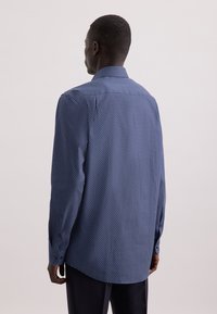 Uomo che indossa una camicia a maniche lunghe blu con pois bianchi, in piedi con la schiena rivolta verso la fotocamera su uno sfondo semplice chiaro.