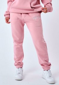 Pantalones de chándal de algodón rosa con puños elásticos, con un logotipo bordado en blanco. Llevados con zapatillas blancas sobre un fondo sencillo.