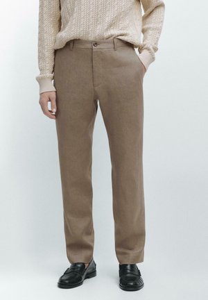 Beiger Strickpullover mit Zopfmuster, kombiniert mit braunen geraden Hosen und schwarzen Leder-Loafers, gezeigt an einer stehenden Person.