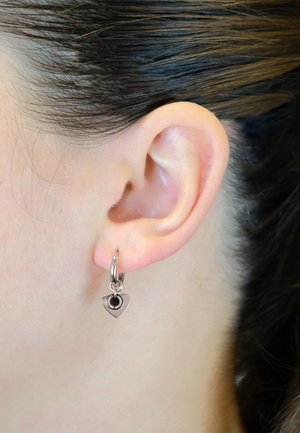 Silberner Creolenohrring mit einem herzförmigen Anhänger, der ein schwarzes Zentrum und filigrane Verzierungen am Rand aufweist, getragen am Ohr einer Person.