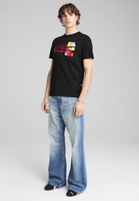 Sort bomulds T-shirt med farverigt geometrisk print; sat sammen med lyseblå flared jeans, båret med sorte sko med hvide logo-detaljer.