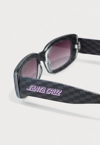 Sorte solbriller med en blank finish, lilla linser og ternet mønster på stængerne med "SANTA CRUZ" i pink skrift.