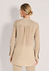 Beige Tunika-Shirt mit langen Ärmeln, glatter Textur, einer Faltenlegung auf der Rückseite und geknöpften Manschetten. Entworfen für eine lockere Passform.