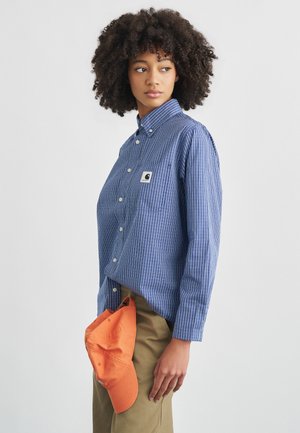 Donna con capelli ricci che indossa una camicia a righe blu abbottonata e pantaloni beige, che tiene un berretto arancione di fianco, guardando di lato.