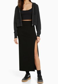 Femme portant un crop hoodie noir, un crop top noir, une longue jupe noire avec une fente haute, des chaussettes noires et des baskets noires avec des semelles en gomme.