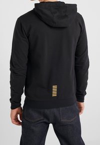 Svart huva sweatshirt med en slät yta, figursydd design och ett horisontellt randigt gulddetalj på baksidan nära fållen.
