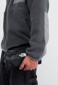 Szürke flaneldzseki világosszürke kiegészítőkkel; bal kézben egy sima felületű és logós fekete The North Face táska. Fekete nadrág.