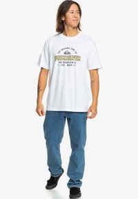 White cotton t-shirt with black and yellow graphic design, paired with blue denim jeans and black sneakers. Casual, loose-fitting style.