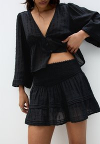 Blusa nera, testurizzata, con scollo a V profondo e chiusura a bottoni, abbinata a una gonna nera a più strati con arricciature. Il tessuto è leggero e fluente.