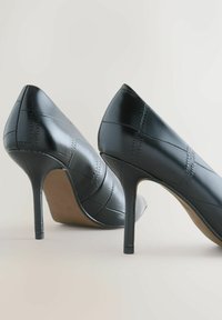 Fekete, hegyes orrú, magas sarkú cipők párja sima bőrön, texturált varrással, egy világos, egyszínű háttér előtt, szögben elhelyezve.