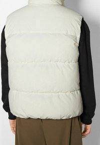 Uppblåsbart väst i ljuskrämfärgad ton, med hög krage, quiltad design och ett mjukt, strukturerat tyg. Bärs över en svart långärmad tröja.