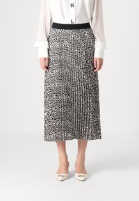 Jupe plissée noir et blanc avec un motif géométrique, en tissu doux, avec une taille élastique. Portée avec des chaussures crème à bout pointu.