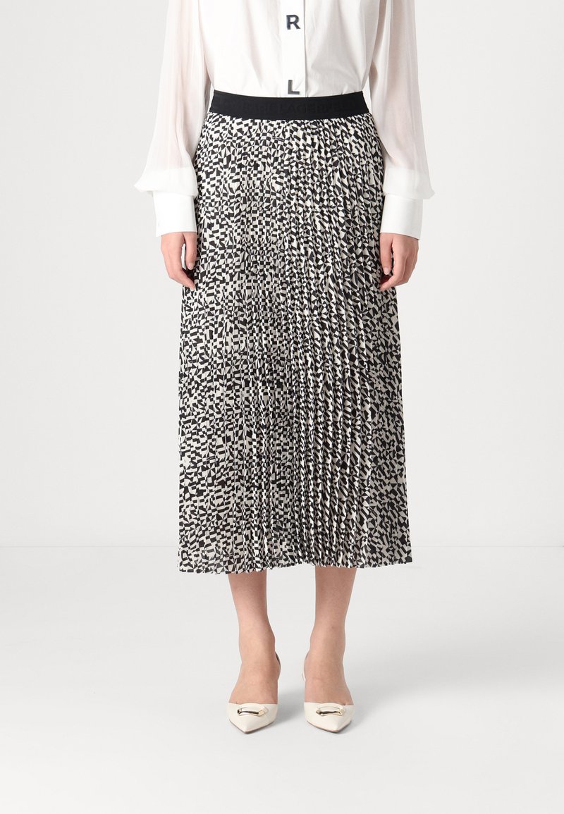 Jupe plissée noir et blanc avec un motif géométrique, en tissu doux, avec une taille élastique. Portée avec des chaussures crème à bout pointu.
