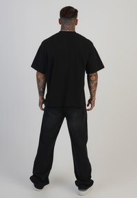 Camiseta negra de manga corta de algodón, combinada con unos pantalones vaqueros oscuros de corte holgado y zapatillas blancas. Tatuajes visibles en los brazos.