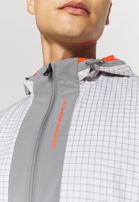 Veste coupe-vent grise à motif quadrillé, avec des accents orange sur la capuche et une fermeture éclair à l'avant. Elle présente un col haut et un tissu léger.