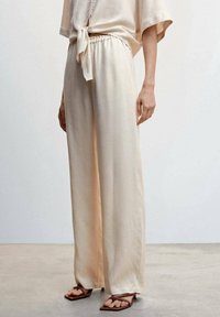 Pantalon beige à jambes larges avec une texture lisse, taille élastique et un dégradé subtil. Associé à des sandales marron.