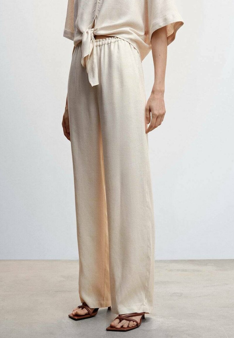 Pantalon beige à jambes larges avec une texture lisse, taille élastique et un dégradé subtil. Associé à des sandales marron.