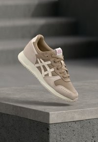 Μπεζ και λευκά sneakers Asics που επιπλέουν πάνω σε μία σκυρόδεμη προεξοχή, με θολές σκάλες στο παρασκήνιο.