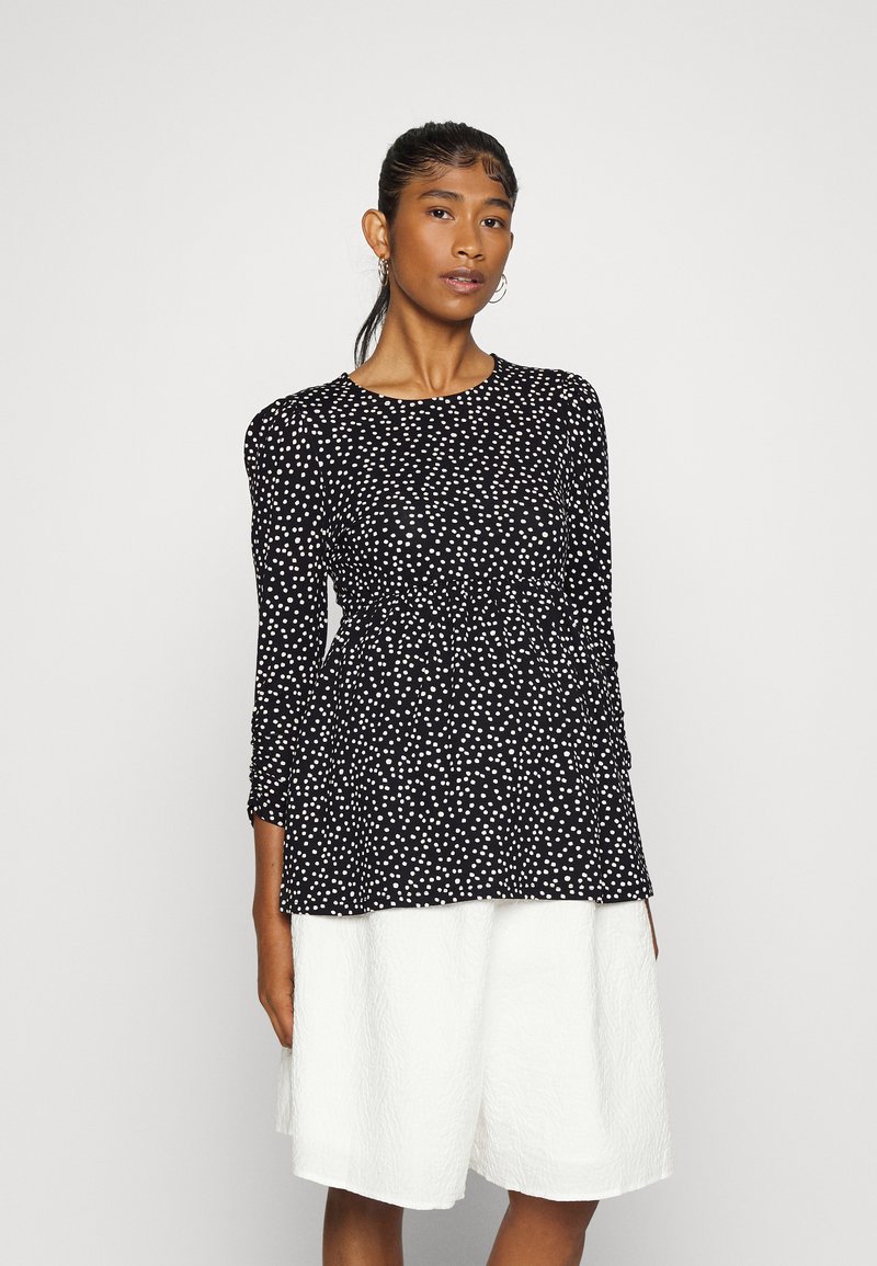 Blusa preta com bolinhas brancas, decote redondo, mangas longas e design peplum, combinada com calções largos brancos texturizados.