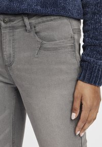 Szürke farmernadrág sima textúrával, első zsebbel, derékpánttal és övhurkakkal, fölötte egy kontrasztos sötétkék mintás pulóver.