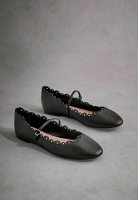 Ballerine in pelle nera con bordi scallop e una cintura decorativa. Punta arrotondata, texture liscia e fodera interna rosa. Suola piatta.