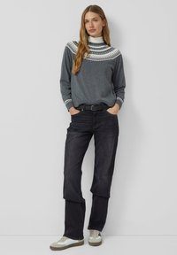 Woman with long hair wearing a gray patterned sweater, white turtleneck, black jeans, black belt, and white sneakers, standing against a light wall.