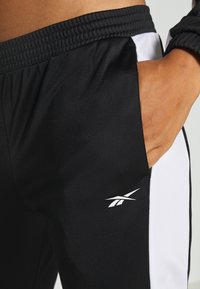Main dans la poche d'un pantalon de sport noir avec une bande latérale blanche et le logo Reebok près de la taille.