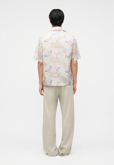 Homme debout, vu de dos, portant une chemise à manches courtes avec un imprimé papillon pastel et un pantalon beige ample sur un fond uni.
