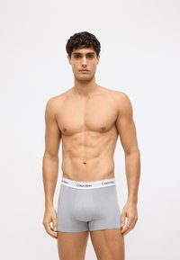 Pelēki stiepjami kokvilnas bokseršortai ar baltu elastīgu jostu, kurā ir izšūts "Calvin Klein" logo, pieguļošs dizains un gluda tekstūra.