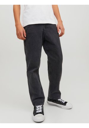 Dolna część ciała osoby noszącej luźne czarne dżinsy i czarno-białe trampki, stojącej na tle jednolitego jasnego tła.